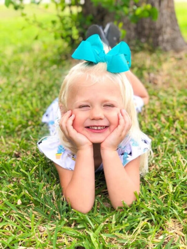 May include: A young child with blonde hair smiles while lying in the grass. The child wears a white dress with a blue floral pattern and a large turquoise bow in their hair. The child rests their chin on their hands.