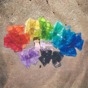 Puede incluir: Una colección de lazos para el cabello coloridos en un espectro de arcoíris, que incluye rojo, naranja, amarillo, verde, azul, morado y negro. Los lazos están dispuestos sobre una superficie texturizada. El texto "Angel Kissed Bows" es visible.