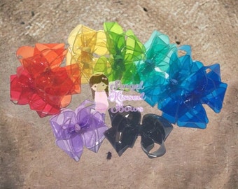May include: A collection of colorful hair bows in a rainbow spectrum, including red, orange, yellow, green, blue, purple, and black. The bows are arranged on a textured surface. The text "Angel Kissed Bows" is visible.