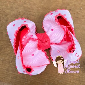 May include: A large, decorative hair bow featuring a layered design with white and pink ribbons. The bow is accented with pink rhinestones and a ruffled pink trim. A smaller, solid pink bow is at the center. The bow has the text "Angel Kissed Bows".