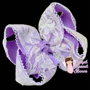 May include: A large, decorative hair bow in shades of lavender and white. The bow features a layered design with a lavender ribbon base, overlaid with white lace and a scalloped edge. A smaller lavender bow is at the center. The background is black.