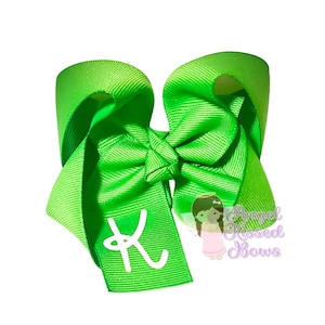 May include: Bright green hair bow with a white letter "K" on the tail. The bow is made of grosgrain ribbon and features a knot in the center. The text "Angel Kissed Bows" is visible in the lower right corner.