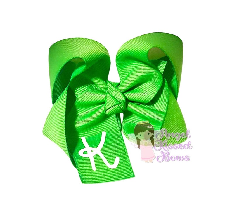 May include: Bright green hair bow with a white letter "K" on the tail. The bow is made of grosgrain ribbon and features a knot in the center. The text "Angel Kissed Bows" is visible in the lower right corner.