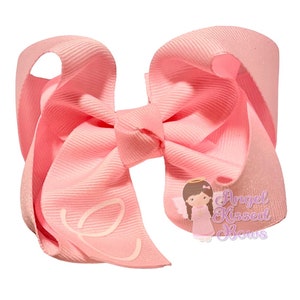 May include: A large, light pink hair bow with a textured grosgrain ribbon. The bow features a stylized white letter "e" on one of the loops. A small illustration of an angel with the text "Angel Kissed Bows" is visible.