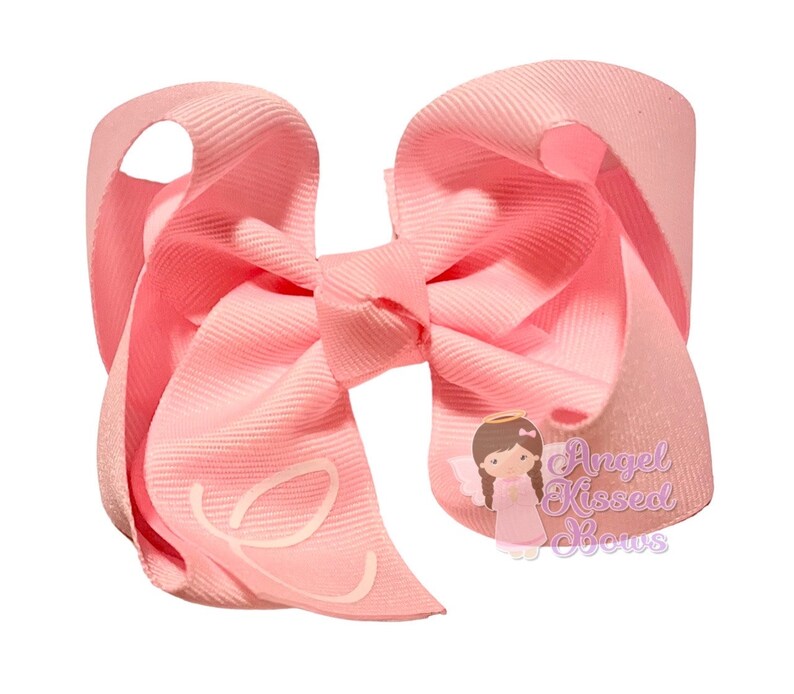 May include: A large, light pink hair bow with a textured grosgrain ribbon. The bow features a stylized white letter "e" on one of the loops. A small illustration of an angel with the text "Angel Kissed Bows" is visible.