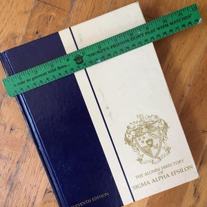 May include: A vintage book, "The Alumni Directory of Sigma Alpha Epsilon," with a navy blue and cream cover. A green ruler with the text "Smokey's Friends Don't Play With Matches" is on top. The book is from 1986.