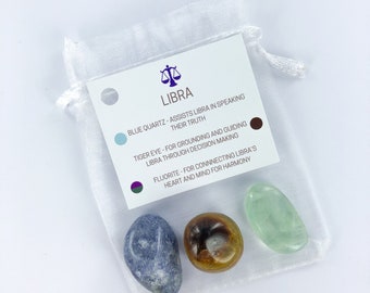 May include: A white organza bag with three crystals inside. The bag has a label that reads "LIBRA" and lists the crystals as blue quartz, tiger eye, and fluorite. The label also describes the properties of each crystal.