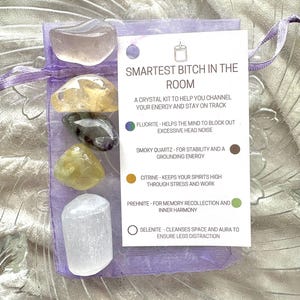 May include: A purple pouch containing a crystal kit with the text "SMARTEST BITCH IN THE ROOM" and a list of crystals and their properties: Fluorite, Smoky Quartz, Citrine, Prehnite, and Selenite.