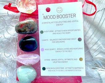 Pode incluir: Uma bolsa vermelha com uma variedade de cristais, incluindo aventurina, unakita, quartzo rosa, citrino e hematita africana. A bolsa tem uma etiqueta branca com o texto "Mood Booster: Um kit de cristais para uma energia positiva e alegre" e descrições de cada cristal.