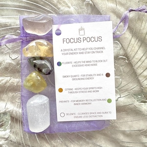 May include: A crystal kit labelled "Focus Pocus" with a purple pouch containing six crystals: clear, yellow, green, brown, and white. The crystals are labelled with their names and properties: Fluorite, Smoky Quartz, Citrine, Prehnite, and Selenite.