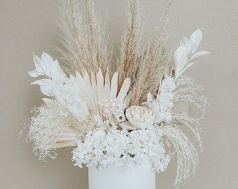 May include: A floral arrangement featuring white dried flowers, pampas grass, and palm spears in a solid white cylindrical vase. The bouquet includes white hydrangea, small white blossoms, and tall, feathery beige pampas grass plumes.