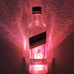 May include: A clear glass bottle with a white label that reads "JOHNNIE WALKER. DOUBLE BLACK." The bottle is lit from the inside with a pink light and is plugged into an electrical outlet.
