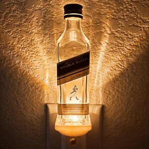 May include: A clear glass bottle with a white label that reads "JOHNNIE WALKER DOUBLE BLACK" and "Johnnie Walker" in a smaller font. The bottle is plugged into a white electrical outlet and is lit from within, creating a warm glow.