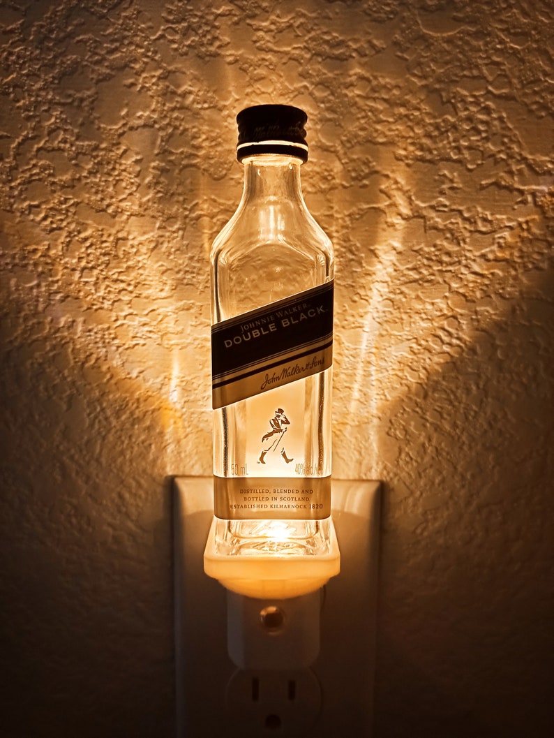 May include: A clear glass bottle with a white label that reads "JOHNNIE WALKER DOUBLE BLACK" and "Johnnie Walker" in a smaller font. The bottle is plugged into a white electrical outlet and is lit from within, creating a warm glow.