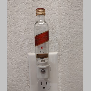 May include: A clear glass bottle with a red label that says "Johnnie Walker Red Label Blended Scotch Whisky" and a picture of a man walking with a cane. The bottle is plugged into a white electrical outlet.