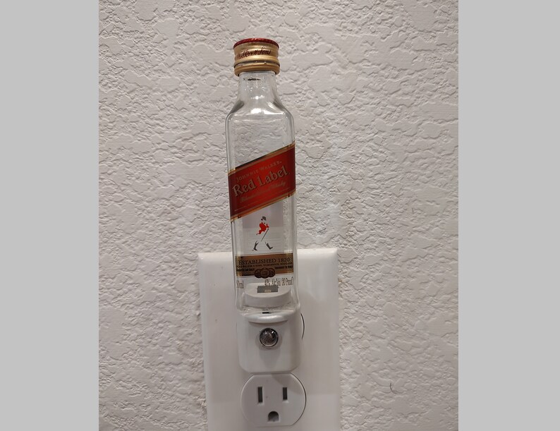May include: A clear glass bottle with a red label that says "Johnnie Walker Red Label Blended Scotch Whisky" and a picture of a man walking with a cane. The bottle is plugged into a white electrical outlet.