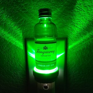 Puede incluir: Una luz nocturna verde con forma de botella de ginebra Tanqueray London Dry. La botella es de vidrio transparente con una etiqueta negra y las palabras "Tanqueray Imported London Dry Gin 47.3% Alc. by Vol. 750 ml" impresas en la etiqueta.
