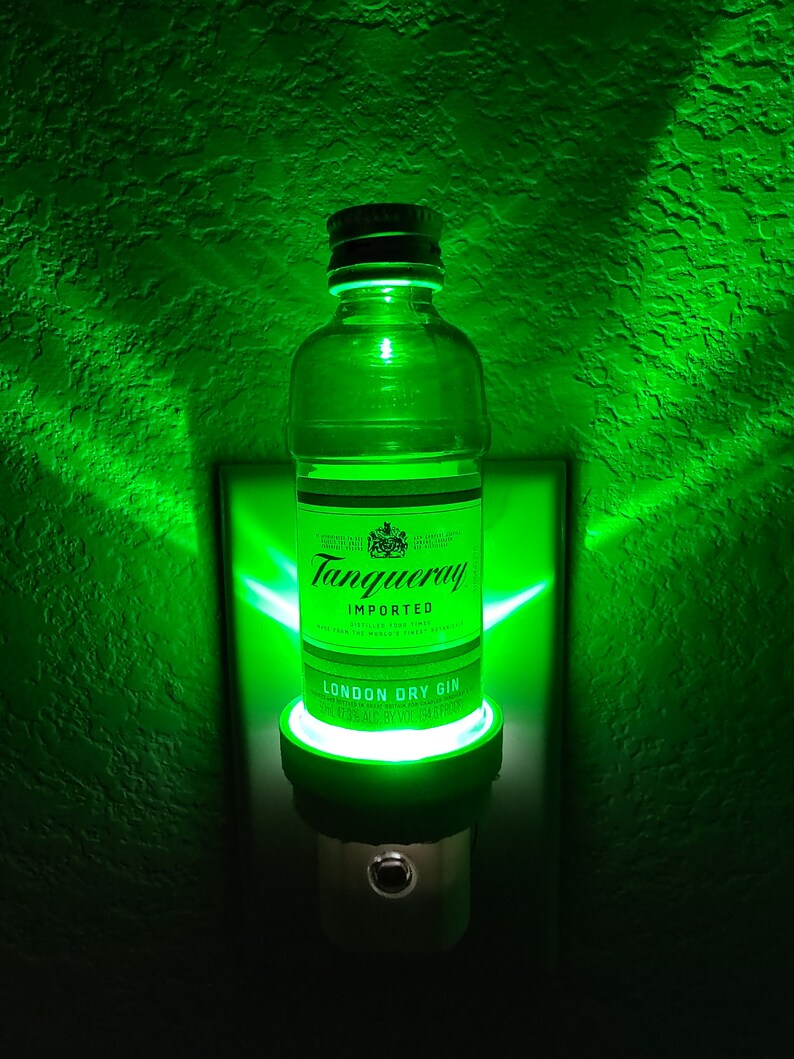Puede incluir: Una luz nocturna verde con forma de botella de ginebra Tanqueray London Dry. La botella es de vidrio transparente con una etiqueta negra y las palabras "Tanqueray Imported London Dry Gin 47.3% Alc. by Vol. 750 ml" impresas en la etiqueta.