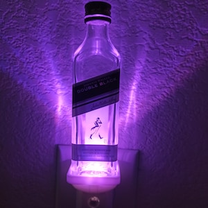 May include: A clear glass bottle nightlight with a white base. The bottle is shaped like a liquor bottle and has a label that reads "Johnnie Walker Double Black" with a silhouette of a man walking with a cane. The bottle is illuminated with a purple light.