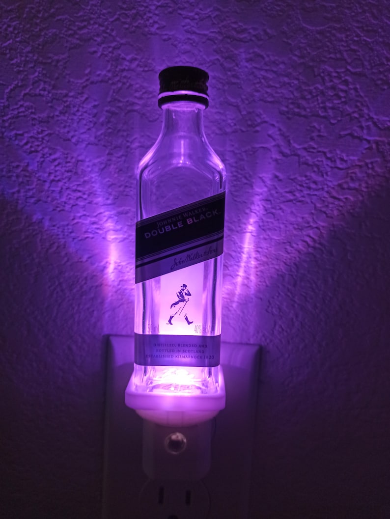 May include: A clear glass bottle nightlight with a white base. The bottle is shaped like a liquor bottle and has a label that reads "Johnnie Walker Double Black" with a silhouette of a man walking with a cane. The bottle is illuminated with a purple light.