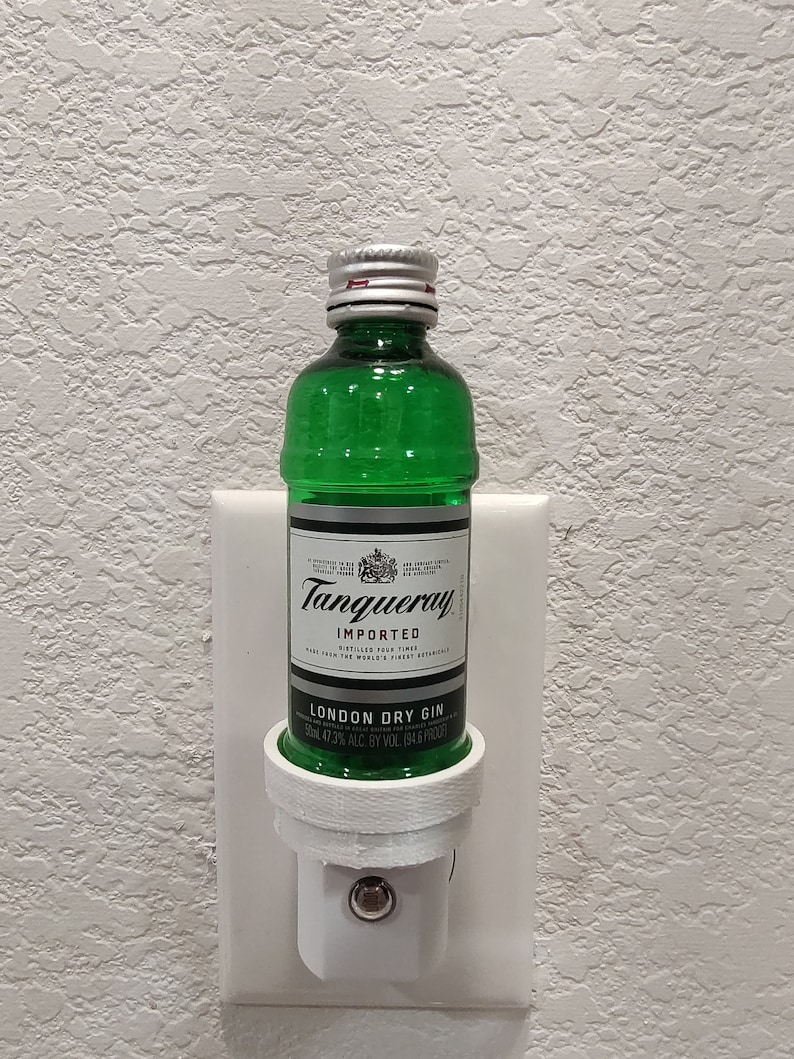 Puede incluir: Una botella de vidrio verde de ginebra seca Tanqueray London Dry Gin, con una tapa plateada, est&aacute; enchufada en una toma de corriente blanca. La botella est&aacute; etiquetada como "Tanqueray Importado" y "Ginebra seca de Londres". La etiqueta tambi&eacute;n indica "47,3% ALC. BY VOL. 194,6 PROOF".