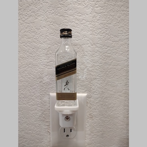 May include: A clear glass bottle with a black label that says "Johnnie Walker Double Black" is plugged into a white nightlight adapter. The bottle is sitting on a white surface.