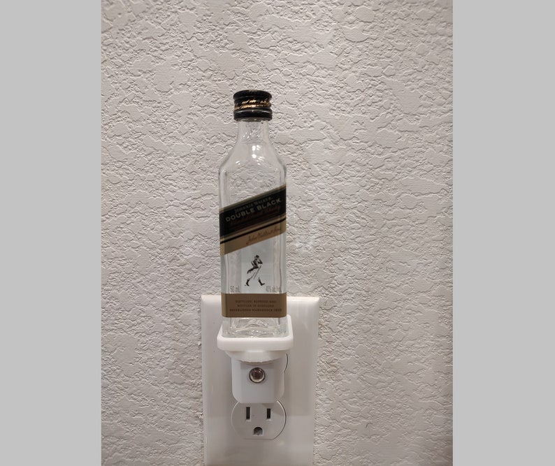 May include: A clear glass bottle with a black label that says "Johnnie Walker Double Black" is plugged into a white nightlight adapter. The bottle is sitting on a white surface.