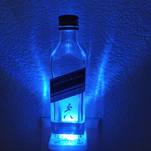 May include: A blue light illuminates a clear glass bottle with a black label that reads "JOHNNIE WALKER DOUBLE BLACK" and features a man with a walking stick. The bottle is plugged into a white electrical outlet.