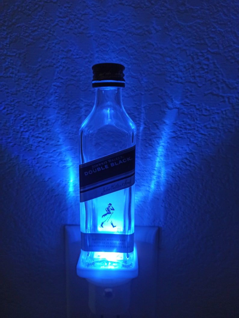 May include: A blue light illuminates a clear glass bottle with a black label that reads "JOHNNIE WALKER DOUBLE BLACK" and features a man with a walking stick. The bottle is plugged into a white electrical outlet.