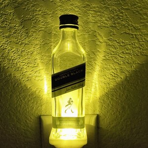 May include: A clear glass bottle night light shaped like a Johnnie Walker Double Black Scotch whisky bottle. The bottle has a black label with white text that reads "Johnnie Walker Double Black" and "Distilled, Blended and Bottled in Scotland. Established Kilmarnock 1820".