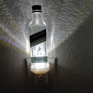May include: A clear glass bottle with a black label that reads "Johnnie Walker Double Black" is plugged into a white electrical outlet. The bottle is lit from inside, creating a warm glow.