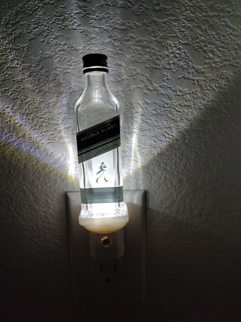 May include: A clear glass bottle with a black label that reads "Johnnie Walker Double Black" is plugged into a white electrical outlet. The bottle is lit from inside, creating a warm glow.