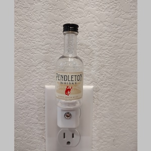 May include: A clear glass bottle with a black cap and a white label that says "Pendleton Whisky" with a red image of a horse. The bottle is plugged into a white electrical outlet.