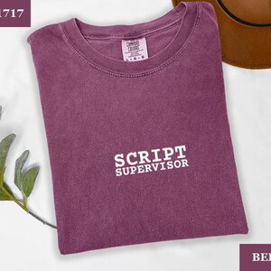 May include: Berry-colored t-shirt with the text "SCRIPT SUPERVISOR" in white block letters. The shirt is a crew neck style and has a tag that says "COMFORT COLORS". The shirt is folded and lying flat.