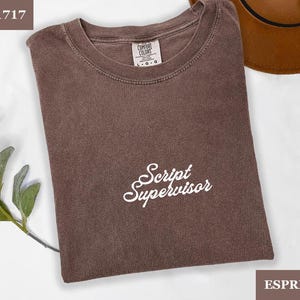 May include: Espresso-coloured t-shirt with the text "Script Supervisor" in white script. The shirt is a Comfort Colors brand, and the size is L-G. A brown hat is visible in the background.