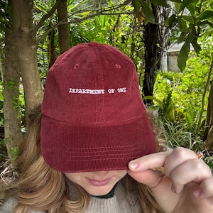 May include: A burgundy corduroy baseball cap with the text "DEPARTMENT OF ONE" embroidered in white. The cap is a classic six-panel design with a curved brim. The background features lush green foliage.