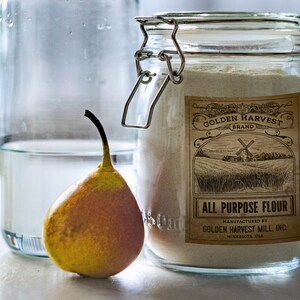 May include: A still life image featuring a glass jar of all-purpose flour with a vintage label, a yellow pear, and a glass bottle with water. The label reads "Golden Harvest Brand" and "All Purpose Flour".