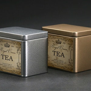 May include: Two metal tea tins, one silver and one gold, with rectangular labels. The labels read "Royal Crown TEA" with ornate scrollwork and the text "Manufactured by Royal Crown Fine Tea Co. London England."