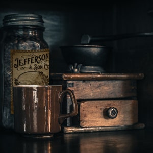 May include: A still life featuring a brown ceramic mug, a glass jar with a label reading "Jefferson & Son Co", and a vintage wooden coffee grinder. The scene is set against a dark background, creating a moody, rustic aesthetic.