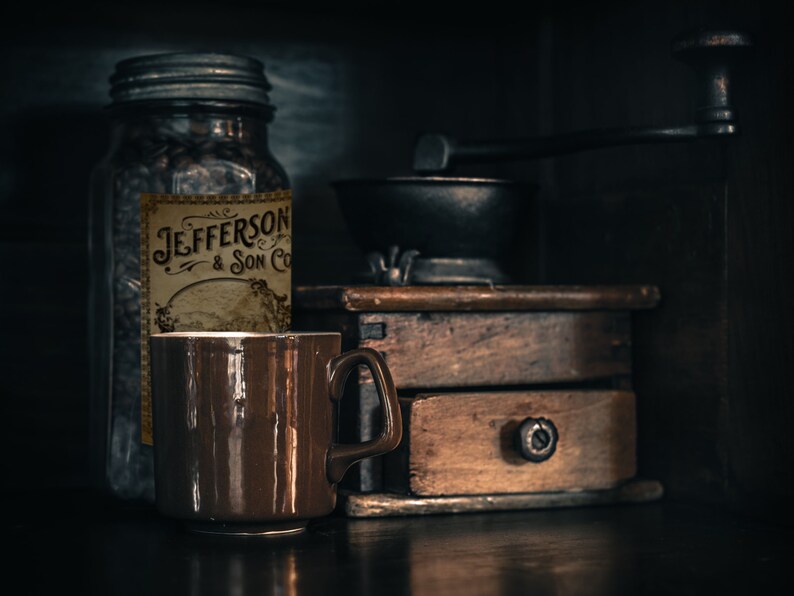 May include: A still life featuring a brown ceramic mug, a glass jar with a label reading "Jefferson & Son Co", and a vintage wooden coffee grinder. The scene is set against a dark background, creating a moody, rustic aesthetic.