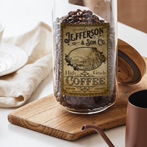 May include: A glass jar filled with coffee beans, labeled "Jefferson & Son Co. High Grade Coffee." The label features an illustration of a landscape. A white teacup and saucer, a wooden spoon with coffee beans, and a brown coffee pot are also visible.