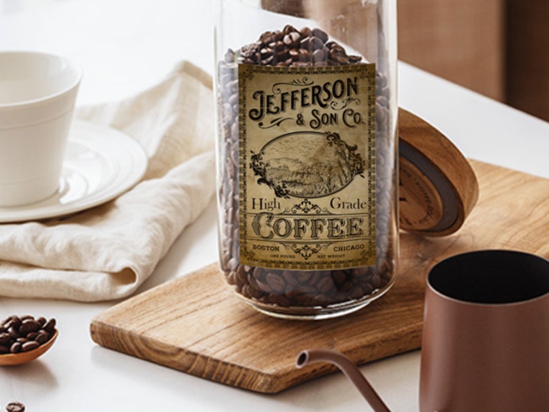 May include: A glass jar filled with coffee beans, labeled "Jefferson & Son Co. High Grade Coffee." The label features an illustration of a landscape. A white teacup and saucer, a wooden spoon with coffee beans, and a brown coffee pot are also visible.