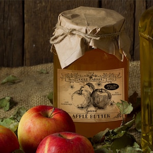 May include: A jar of spiced apple butter with a vintage label featuring apple illustrations. The jar is sealed with brown paper and twine, surrounded by red apples and green leaves. The label reads "Gold Barrel 1879".