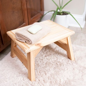 May include: A wooden step stool with a light brown towel and a bar of soap on top. The stool is sitting on a light brown rug.