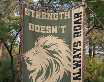 May include: A green and tan woven throw blanket with the text "STRENGTH DOESN'T ROAR ALWAYS ROAR" and a lion's head graphic. The blanket is hanging from a clothesline with wooden clothespins.