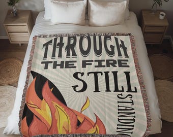 May include: A woven throw blanket with the words "THROUGH THE FIRE STILL STANDING" in black lettering. The blanket features a graphic of flames in shades of red, orange, and yellow. The background is a light green with radiating lines.
