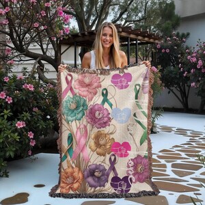 May include: A beige throw blanket with a floral and ribbon design. The blanket features various flowers in shades of pink, teal, purple, and brown, along with ribbons and hearts. The word "love" is also visible. The blanket has a fringed edge.