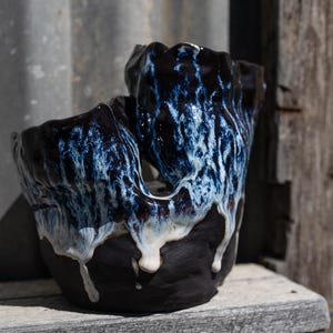 May include: A handmade ceramic bowl with an unusual, organic form. The bowl has a dark, matte black base with a glossy, dripped glaze in shades of blue and white. The bowl is displayed on a weathered wooden shelf.