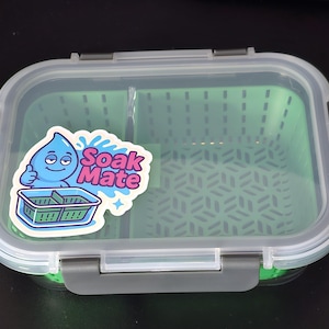 May include: A clear, rectangular food container with a green interior and a clear lid. The container has a sticker that says "Soak Mate" with a cartoon water droplet giving a thumbs up. The container has grey latches.