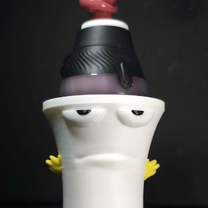 May include: A white, 3D-printed character with a black top, purple base, and red stem. The character has yellow hands and a grumpy expression. The design is a novelty item.